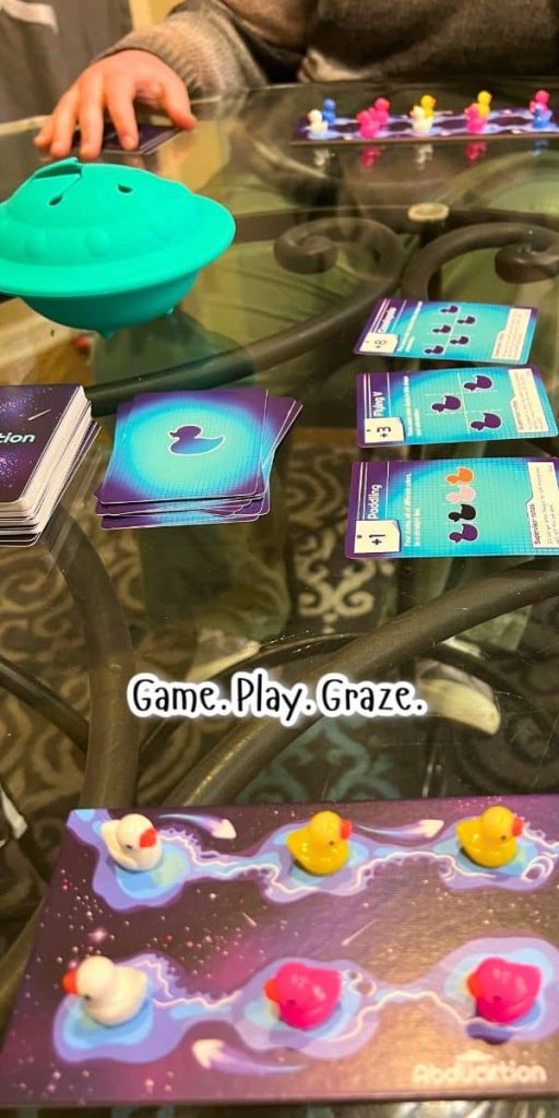 The Duck Board Game for Fun Family Game Nights The Duck Family Board Game - hands playing the Abduction game on a table with mini game boards, colorful ducks, action cards and plastic UFO